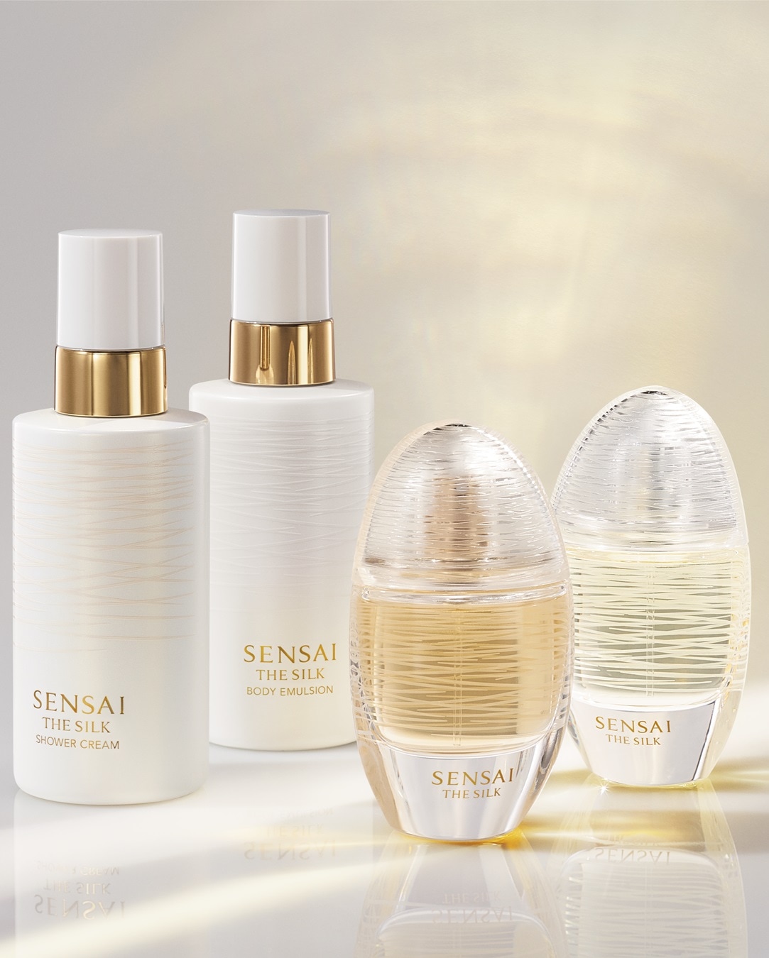 Frascos brancos de creme de banho e emulsão corporal Sensai The Silk, com detalhes dourados, ao lado de frascos ovais transparentes do mesmo perfume.