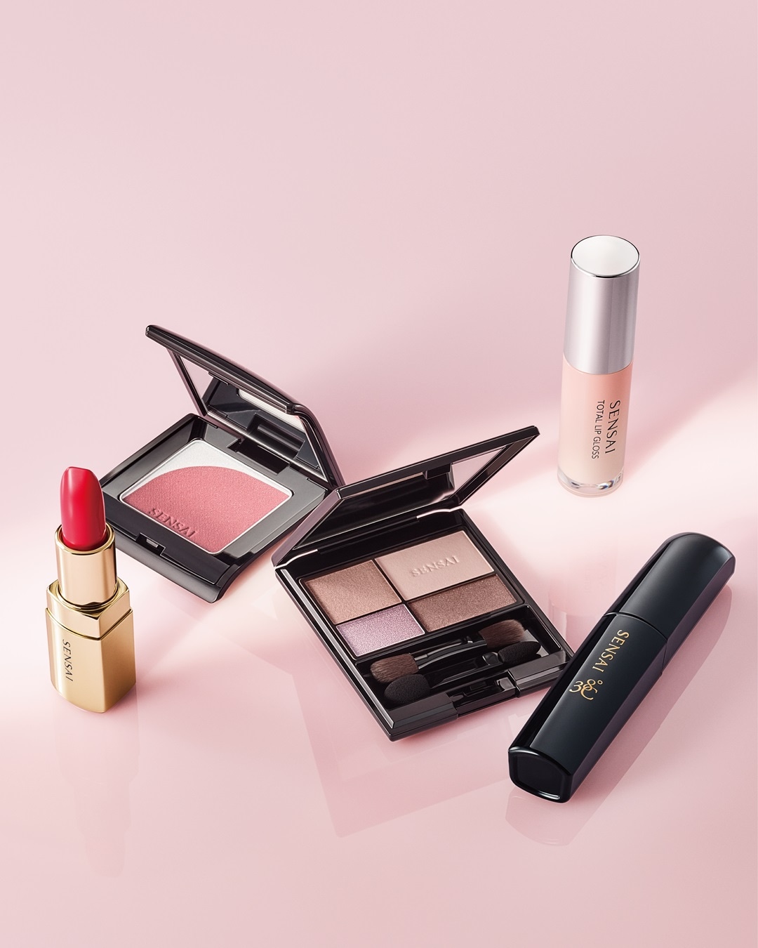 Na różowym tle znajdują się produkty do makijażu Sensai, w tym szminka w złotym opakowaniu, róż do policzków, paleta cieni do powiek, błyszczyk do ust i tusz do rzęs w czarnym opakowaniu.