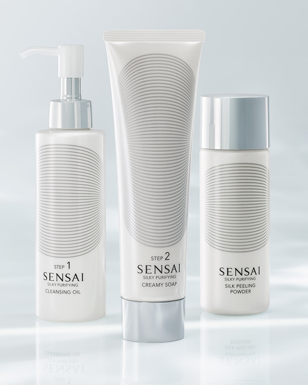 Conjunto de produtos de limpeza facial Sensai Silky Purifying, incluindo óleo de limpeza, sabonete cremoso e pó peeling de seda, todos em embalagens brancas com detalhes prateados.