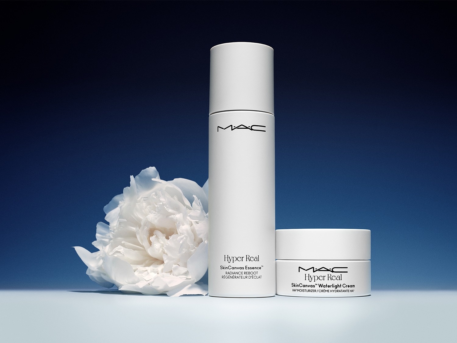 De MAC Hyper Real SkinCanvas Essence en SkinCanvas Waterlight Cream, beide in strakke witte verpakkingen, staan naast een delicate witte bloem tegen een diepblauwe achtergrond.
