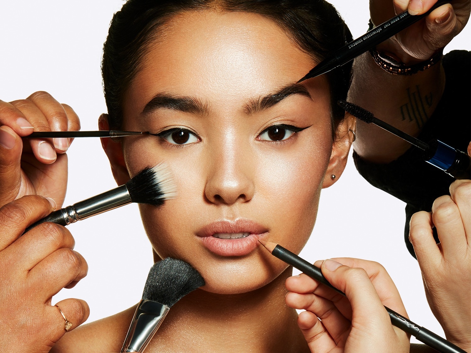 Een close-up van een vrouw die make-up krijgt, met meerdere handen die verschillende make-upborstels en producten vasthouden, zoals eyeliner, mascara, lipliner en poederkwasten, rond haar gezicht.