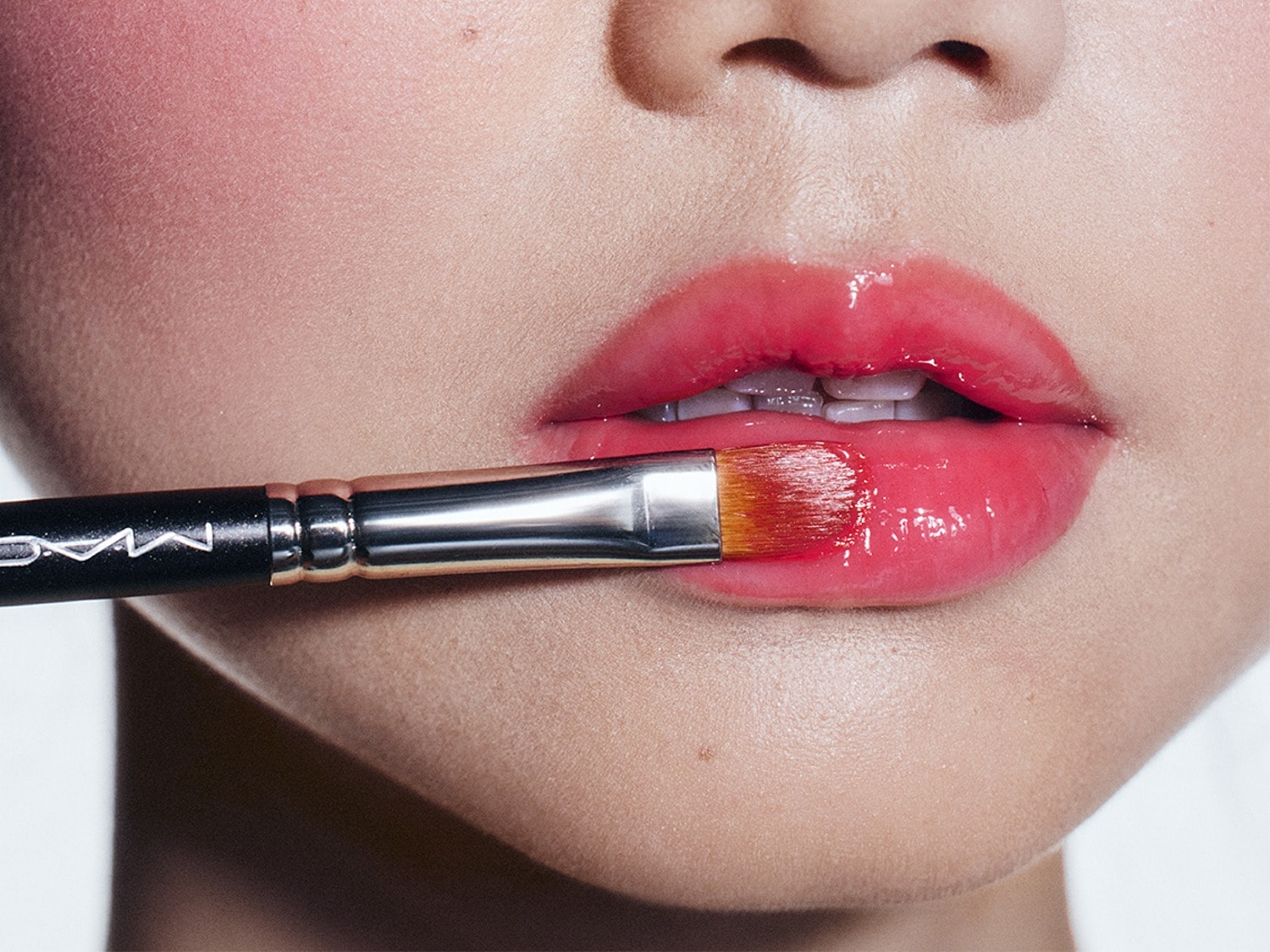 Een close-up van een gezicht toont een MAC-kwast die een glanzende, koraalroze lipgloss aanbrengt op de lippen, met een subtiele blos op de wangen.