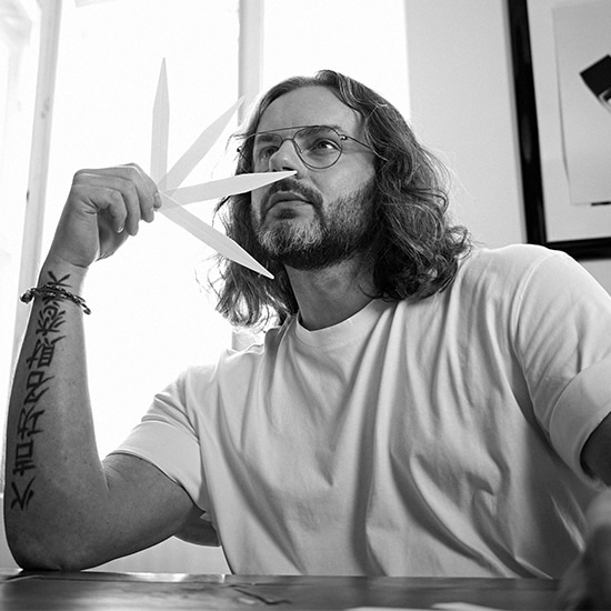 Czarno-białe zdjęcie mężczyzny z długimi włosami i brodą, który trzyma wachlarz białych pasków pod nosem, prawdopodobnie wąchając perfumy.