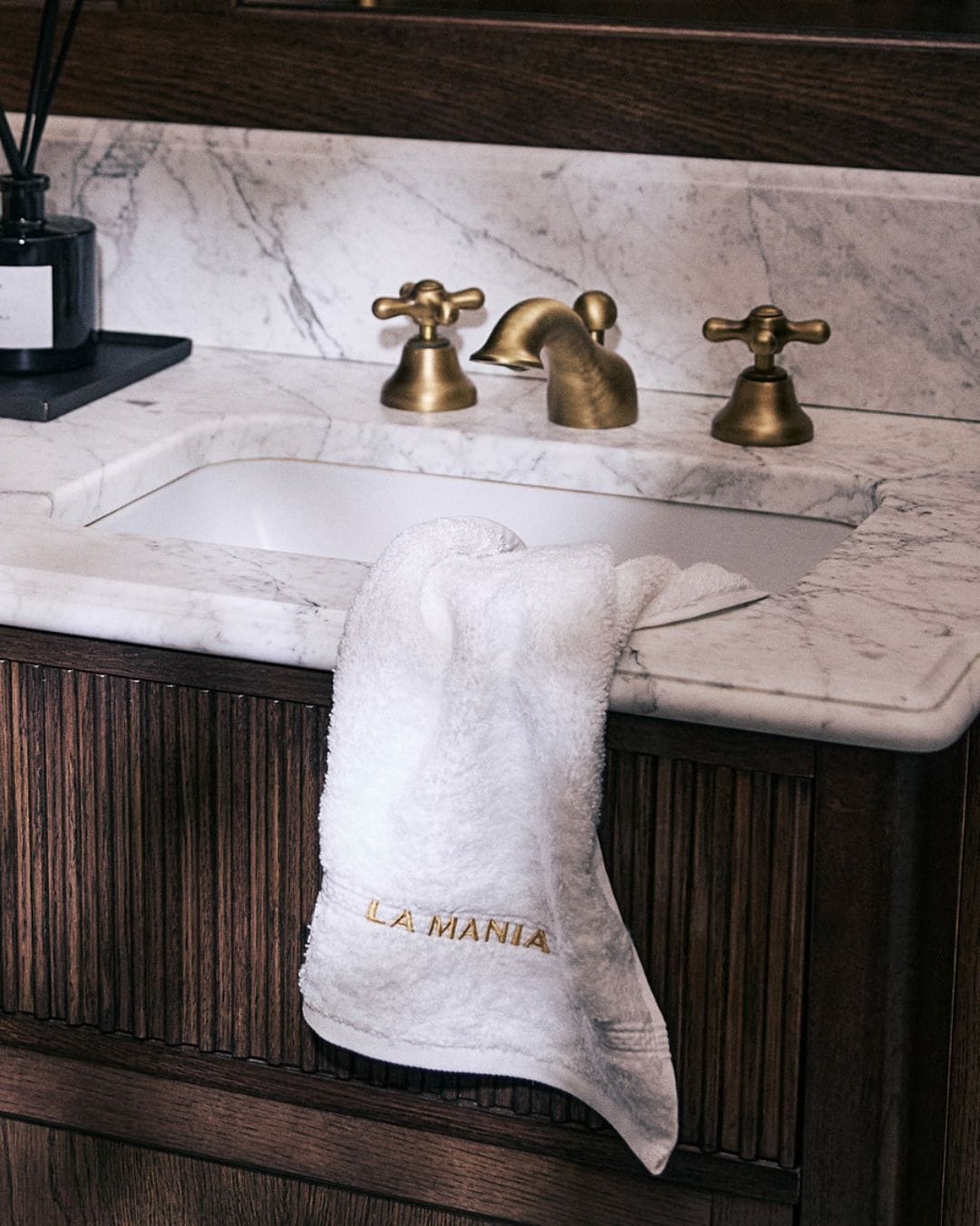 Na marmurowym blacie umywalki leży biały ręcznik z wyhaftowanym złotym napisem "LA MANIA", obok stoi czarny dyfuzor zapachowy, a całość dopełnia bateria umywalkowa w kolorze starego złota.
