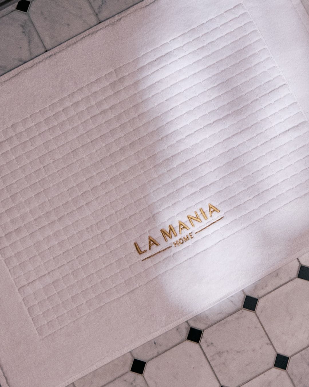 Biały, prostokątny dywanik łazienkowy z napisem „LA MANIA HOME” w kolorze złotym, leżący na marmurowej podłodze.