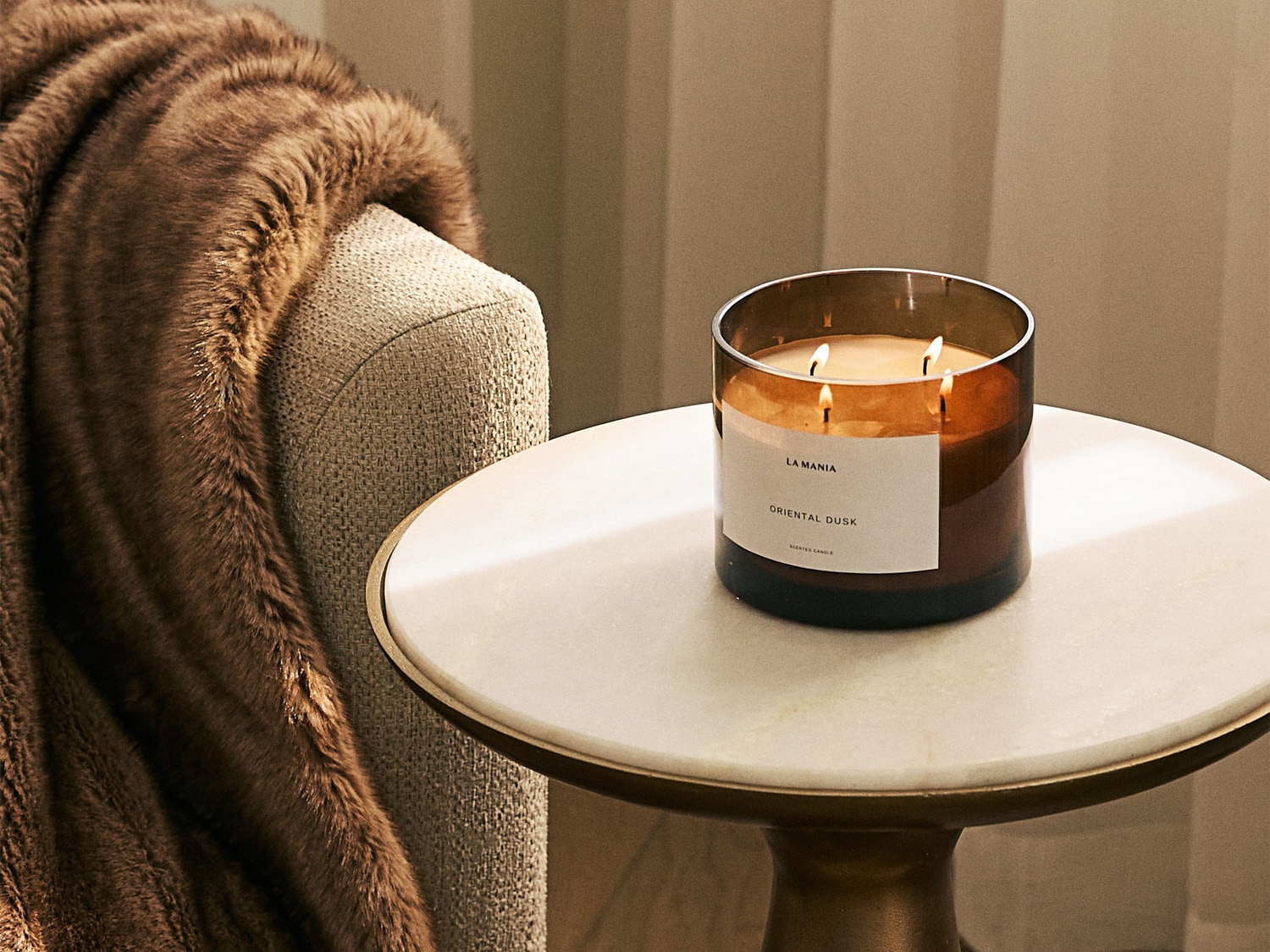 Na stoliku nocnym stoi zapalona świeca La Mania Oriental Dusk w szklanym, brązowym pojemniku, obok leży miękki, brązowy koc.