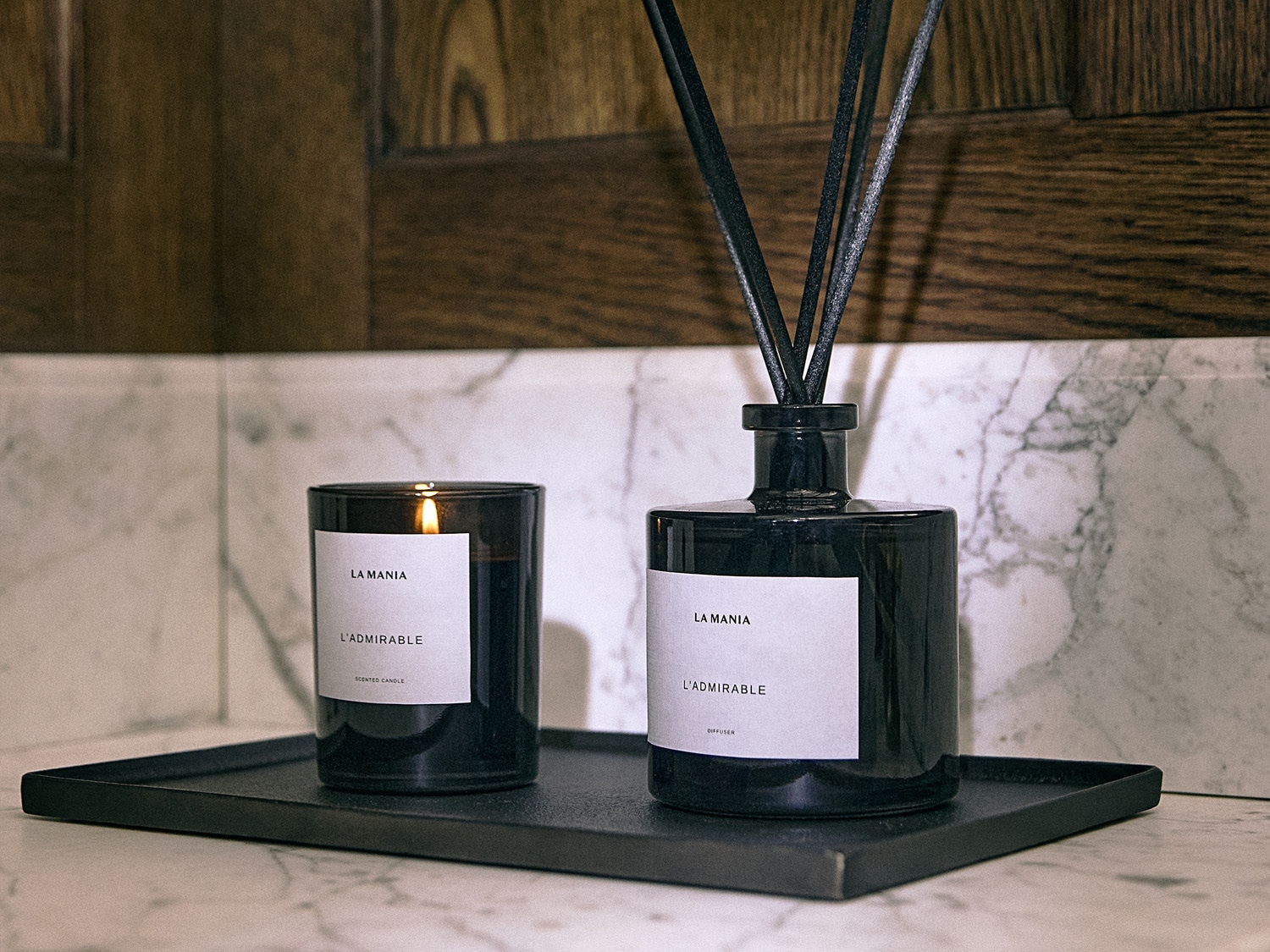 Czarny świeca zapachowa La Mania L'Admirable i dyfuzor z czarnymi patyczkami na czarnej tacy, tworzące elegancki zestaw do aromaterapii.