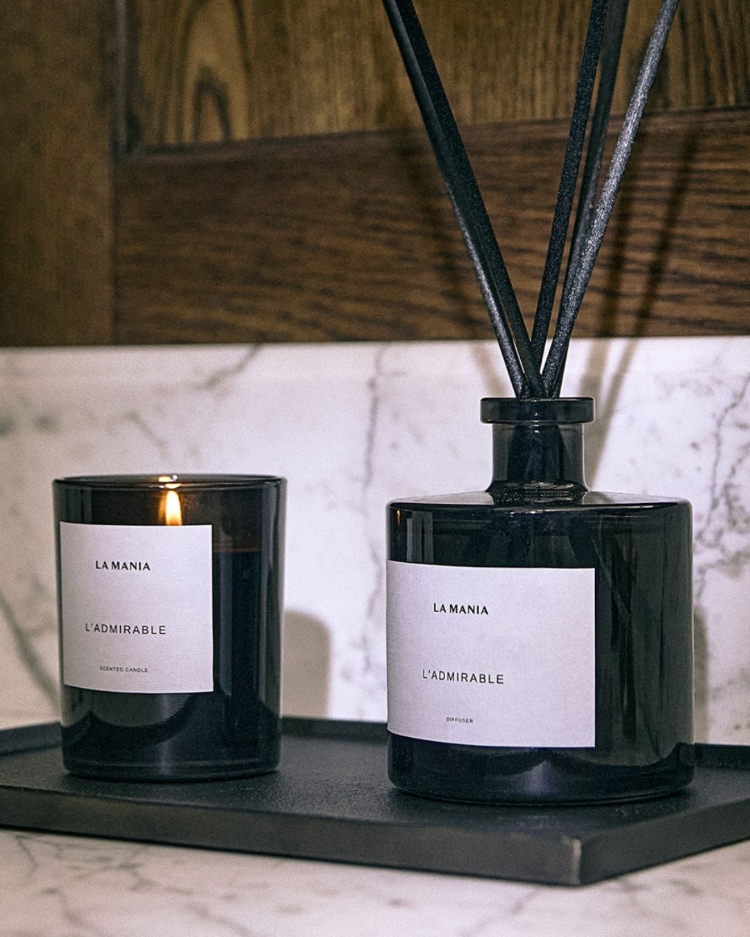 Czarny świeca zapachowa La Mania L'Admirable obok dyfuzora zapachowego z czarnymi patyczkami, oba na czarnej tacy, tworzące elegancki zestaw do aromaterapii.
