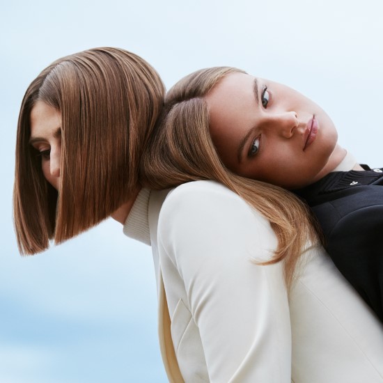 Dwie kobiety opierają się plecami o siebie, jedna ma krótkie brązowe włosy i białą marynarkę, a druga długie blond włosy i czarną marynarkę.