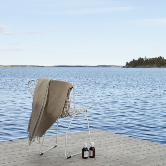 Na drewnianym pomoście, przy białym krześle z beżowym ręcznikiem, stoją dwie brązowe butelki kosmetyków na tle błękitnego jeziora.