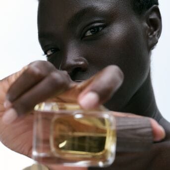 Nahaufnahme einer dunkelhäutigen Frau, die ein Parfüm mit goldenem Verschluss in der Hand hält.