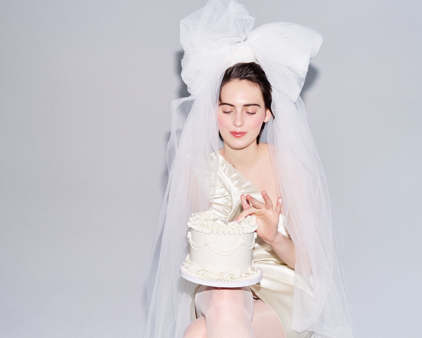 Una donna vestita di bianco con un grande fiocco e un velo, tiene in mano una torta bianca decorata, con gli occhi chiusi e un'espressione serena.