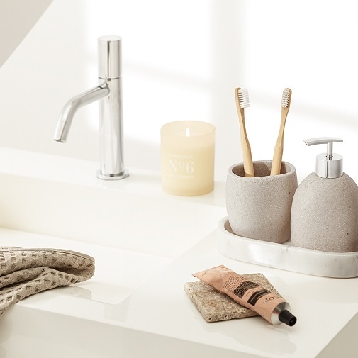 Na blacie umywalki stoi zapalona świeca, dozownik do mydła, kubek ze szczoteczkami do zębów, krem do rąk i ręcznik.