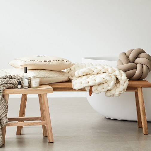 Na drewnianej ławce leżą beżowe poduszki, biały koc z grubym splotem i dekoracyjny węzeł, obok stoi świeca i flakonik z zapachem.