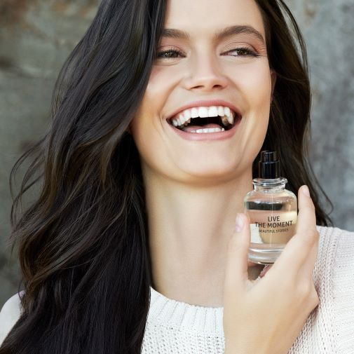 Eine strahlende Frau hält ein Parfümfläschchen mit der Aufschrift „Live the Moment Beautiful Stories“ in der Hand.