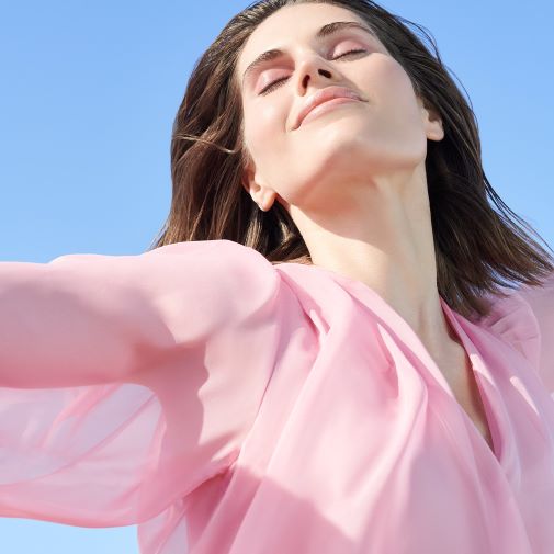 Eine Frau mit geschlossenen Augen und einem rosa Oberteil genießt die Sonne unter einem strahlend blauen Himmel.