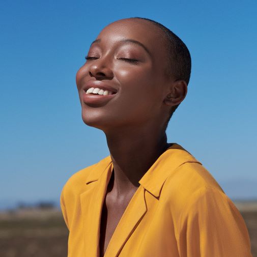 Eine Frau mit dunkler Haut und kurz geschorenen Haaren genießt die Sonne mit geschlossenen Augen und einem strahlenden Lächeln, gekleidet in eine gelbe Jacke vor einem strahlend blauen Himmel.