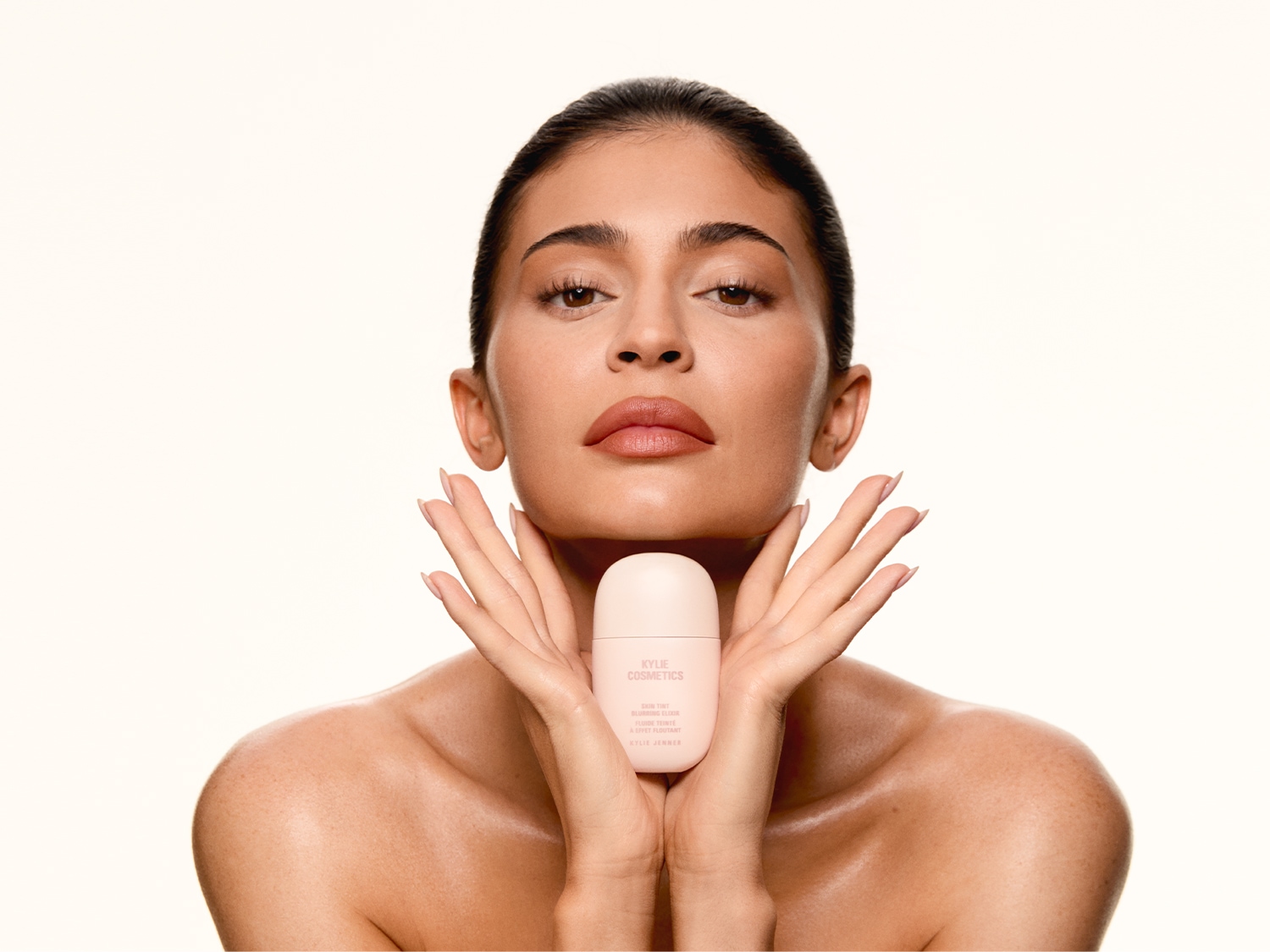 Kobieta z nagimi ramionami trzyma w dłoniach jasnoróżową butelkę Kylie Cosmetics Skin Tint, prezentując produkt do makijażu.