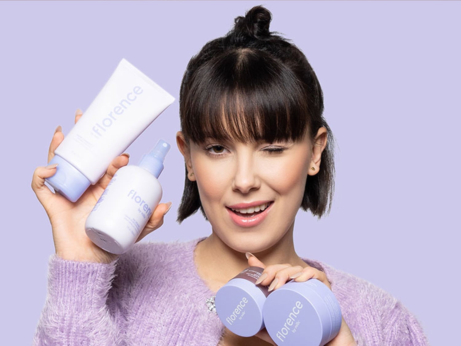 Młoda kobieta z krótkimi brązowymi włosami i grzywką, uśmiecha się i mruga okiem, prezentując serię produktów do pielęgnacji skóry Florence by Mills w białych i liliowych opakowaniach.