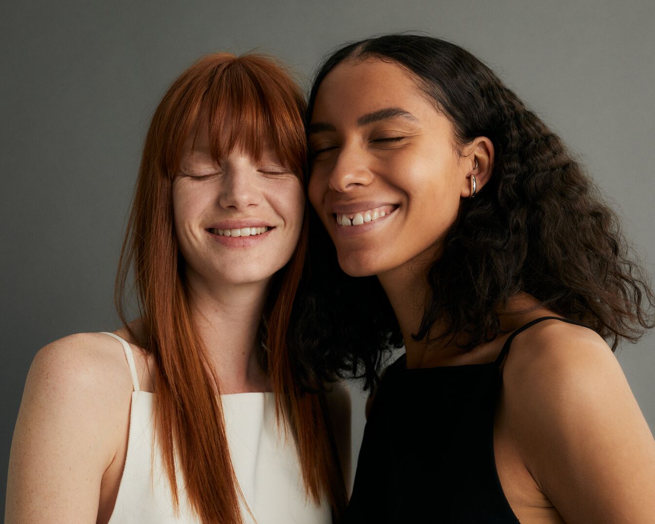 Dos mujeres sonriendo con los ojos cerrados, una con el pelo liso rojizo y la otra con el pelo rizado oscuro, ambas con camisetas de tirantes.