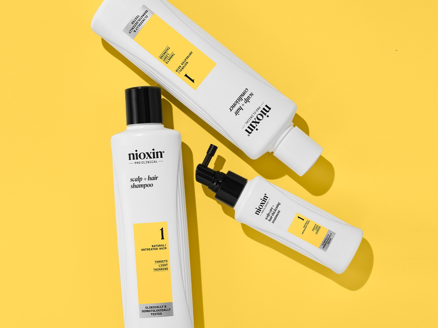 Na imagem, três produtos Nioxin Pro-Clinical contra queda de cabelo: shampoo, condicionador e tratamento, todos em embalagens brancas com detalhes em amarelo e preto, sobre um fundo amarelo.
