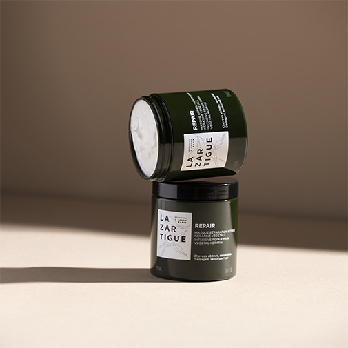 Dwa słoiki z maską do włosów LAZARTIGUE Repair, jeden na drugim, na beżowym tle. Maska ma ciemnozielone opakowanie z białą etykietą z napisem REPAIR i nazwą marki.