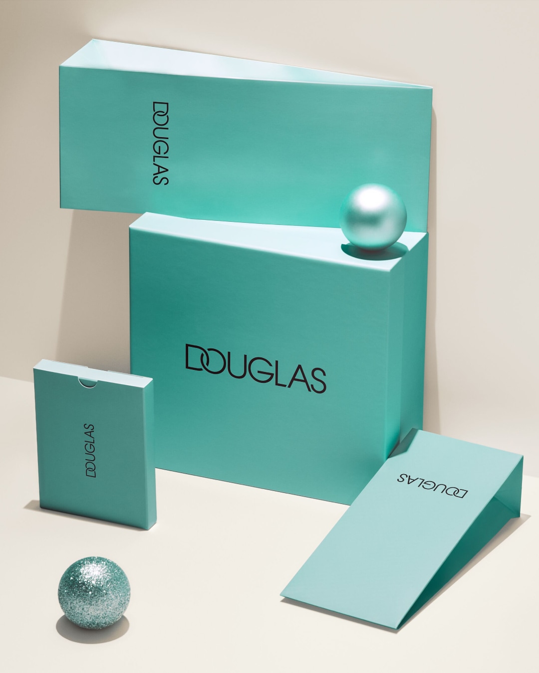 Na kremowym tle znajdują się turkusowe pudełka Douglas o różnych rozmiarach, ułożone jeden na drugim, a obok nich leżąca błyszcząca kula.