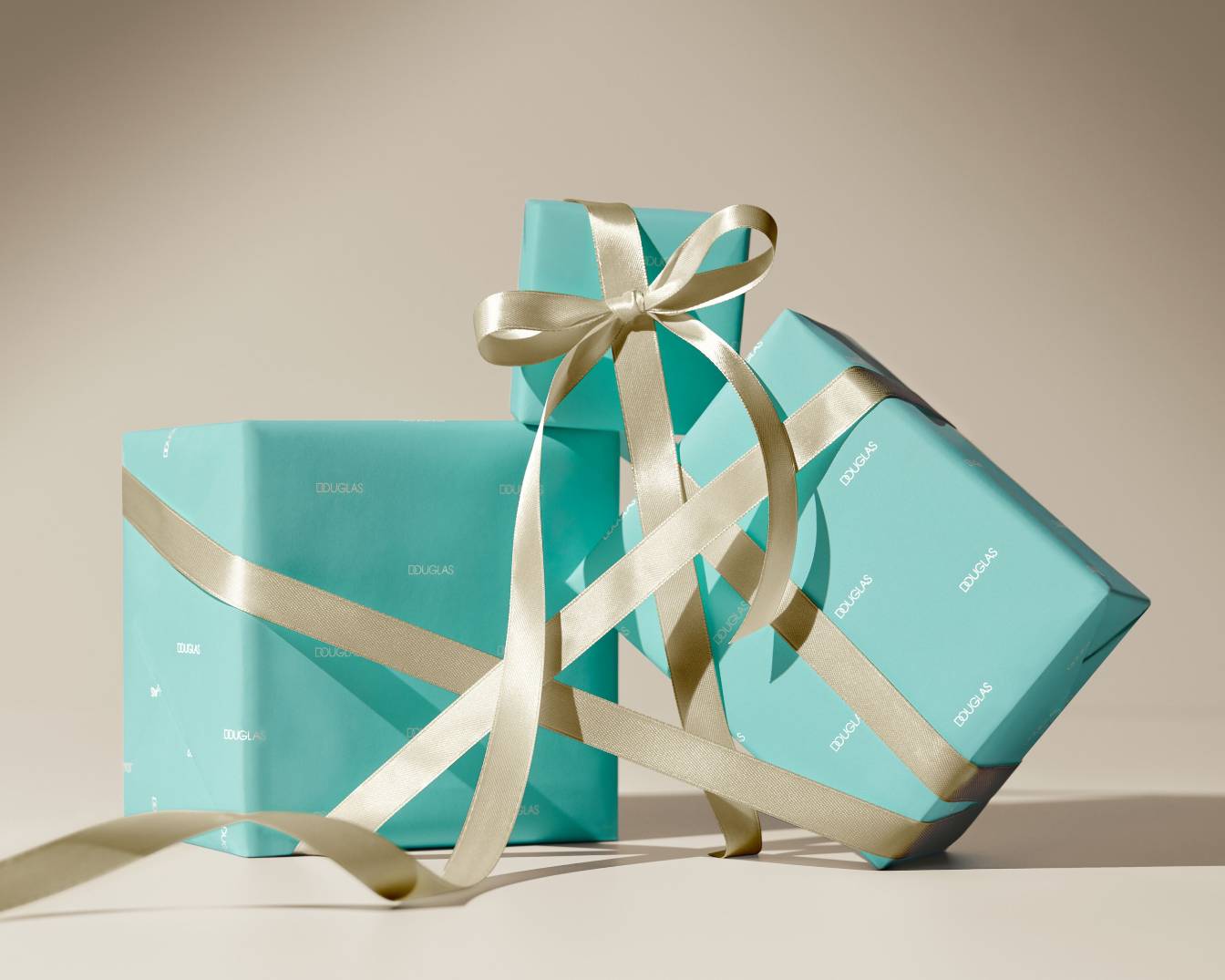 Trzy prezenty Douglas owinięte w turkusowy papier z logo i przewiązane złotą wstążką, tworzące elegancką kompozycję.