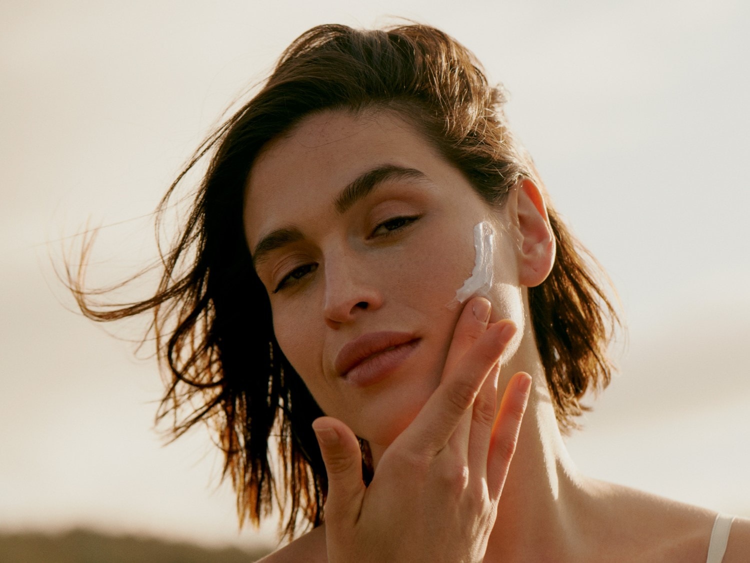 Eine Frau mit braunen Haaren trägt eine weiße Creme auf ihre Wange auf.
