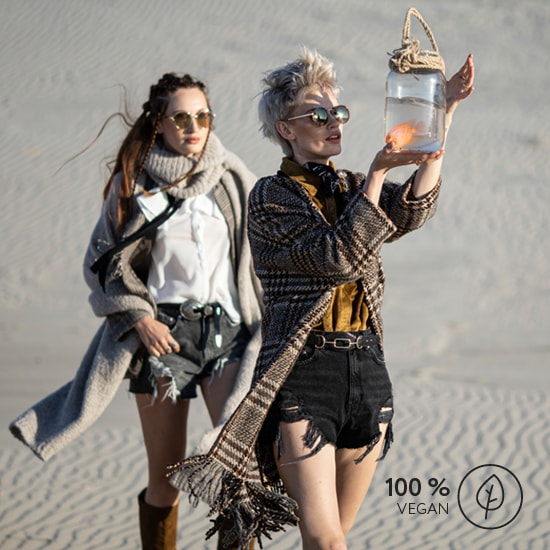 Dwie kobiety w okularach przeciwsłonecznych stoją na pustyni, jedna trzyma słoik ze złotą rybką, a w prawym dolnym rogu znajduje się logo 100% Vegan.