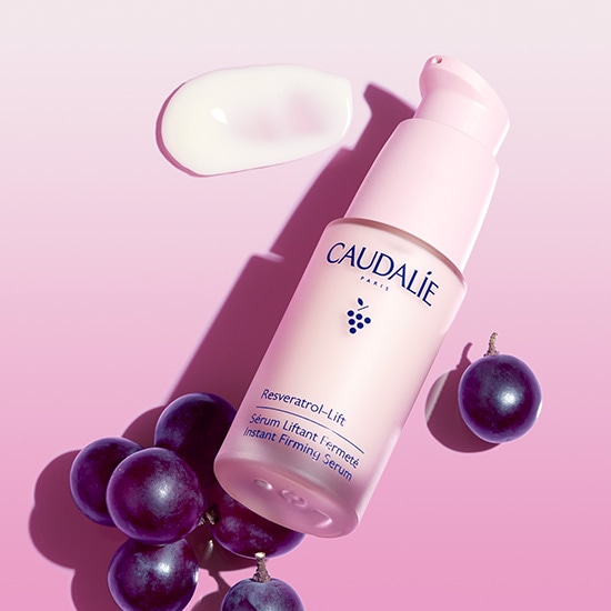 Różowe serum Caudalie Resveratrol-Lift Instant Firming Serum z ekstraktem z winogron, obok leży próbka kremu i kilka winogron.