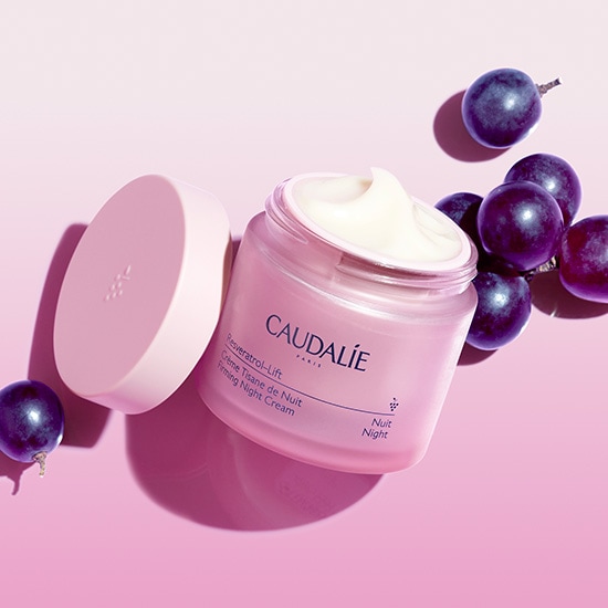 Różowy słoiczek kremu Caudalie Resveratrol-Lift Firmin Night Cream z otwartym wieczkiem, obok leżą ciemne winogrona na różowym tle.