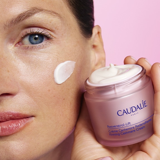 Kobieta nakłada krem Caudalie Resveratrol-Lift na twarz z otwartego słoiczka w różowym opakowaniu, prezentując jego aksamitną konsystencję.