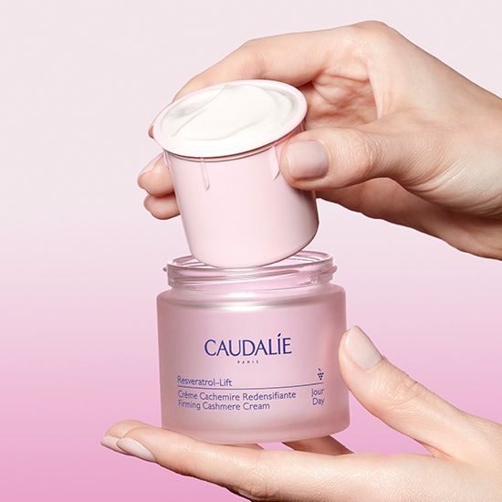 Ręce trzymają różowy słoiczek kremu Caudalie Resveratrol-Lift Crème Cachemire Redensifiante Firming Cashmere Cream z otwartym wieczkiem, ukazując jego aksamitną konsystencję.