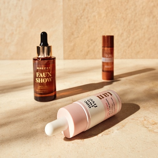 Na pierwszym planie leży buteleczka One Two Free Tan Drops z białą pipetą i różową nakrętką, w tle widać Morphe Faux Show w brązowej butelce i Sun Face Stick w pomarańczowym opakowaniu.