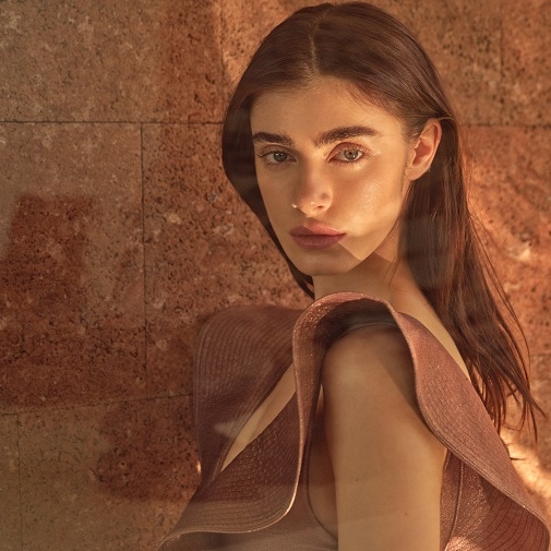 Portret kobiety z długimi, brązowymi włosami i delikatnym makijażem, ubrana w sukienkę z dużym, falującym kołnierzem w odcieniach brązu, pozuje na tle ściany z cegieł.