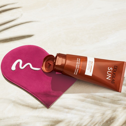 Brązowa tubka samoopalacza Douglas Sun leży obok różowej rękawicy do aplikacji, na którą wyciśnięto biały krem, zapewniając stopniową opaleniznę twarzy i ciała z efektem naturalnego blasku.