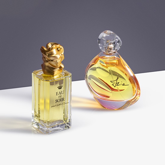 Dwa flakony perfum: Eau du Soir w prostokątnym flakonie ze złotą nakrętką w kształcie popiersia i Izïa w zaokrąglonym flakonie z przezroczystą nakrętką, oba wypełnione płynem o ciepłym, żółtym odcieniu.