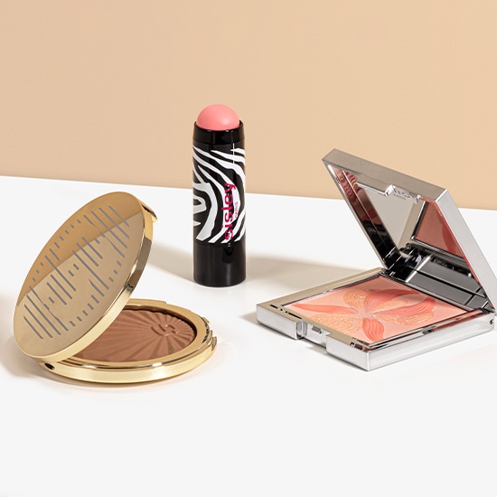 Na białym tle leżą trzy produkty do makijażu: bronzer w złotym opakowaniu, róż w sztyfcie w czarno-białe paski zebry i rozświetlacz w srebrnym opakowaniu.