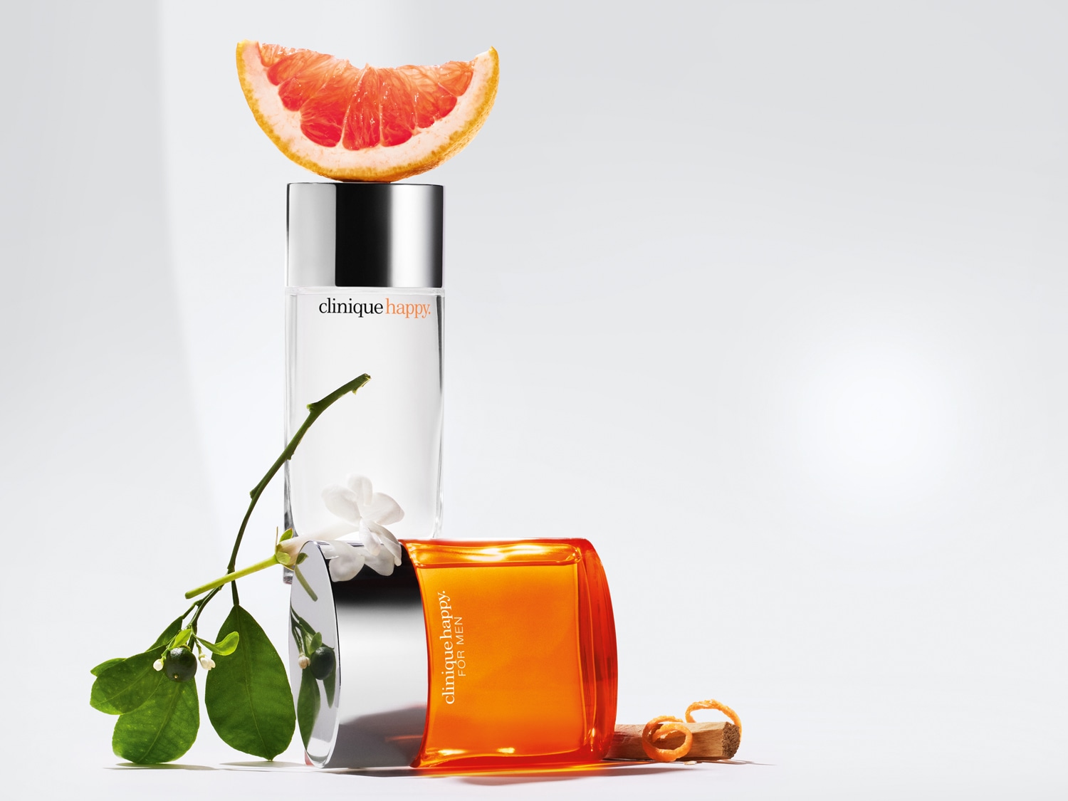 Twee flessen Clinique Happy parfum, de een helder en de ander oranje, staan naast een takje met witte bloemen en een schijfje grapefruit.