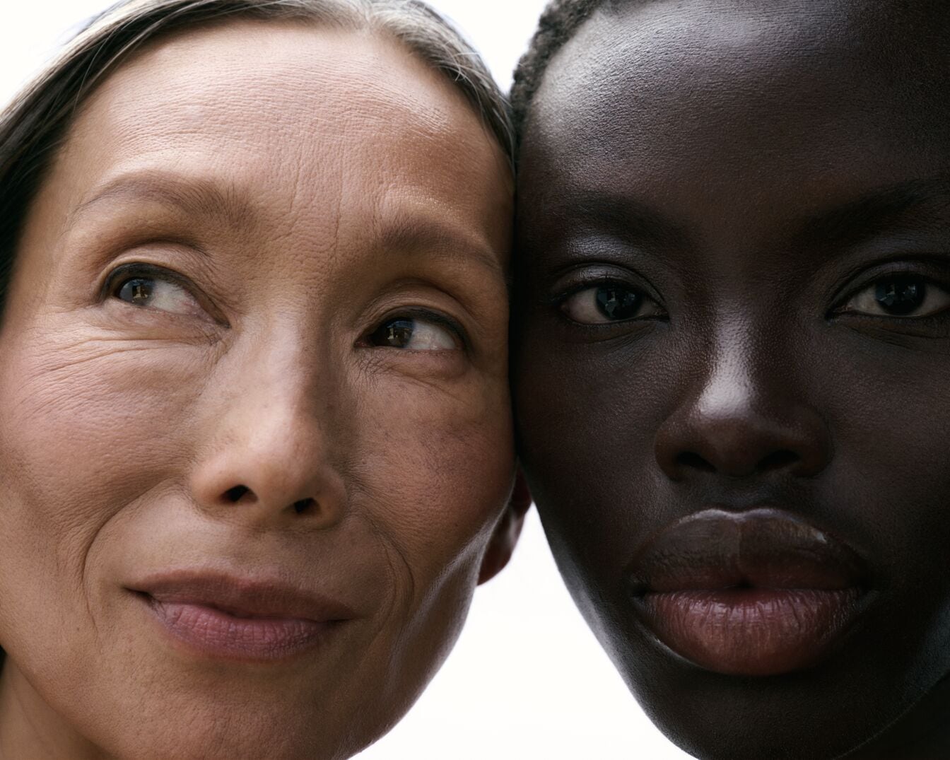 Šalia viena kitos stovi dvi moterys, viena su šviesia oda, o kita su tamsia oda, žiūrinčios skirtingomis kryptimis.