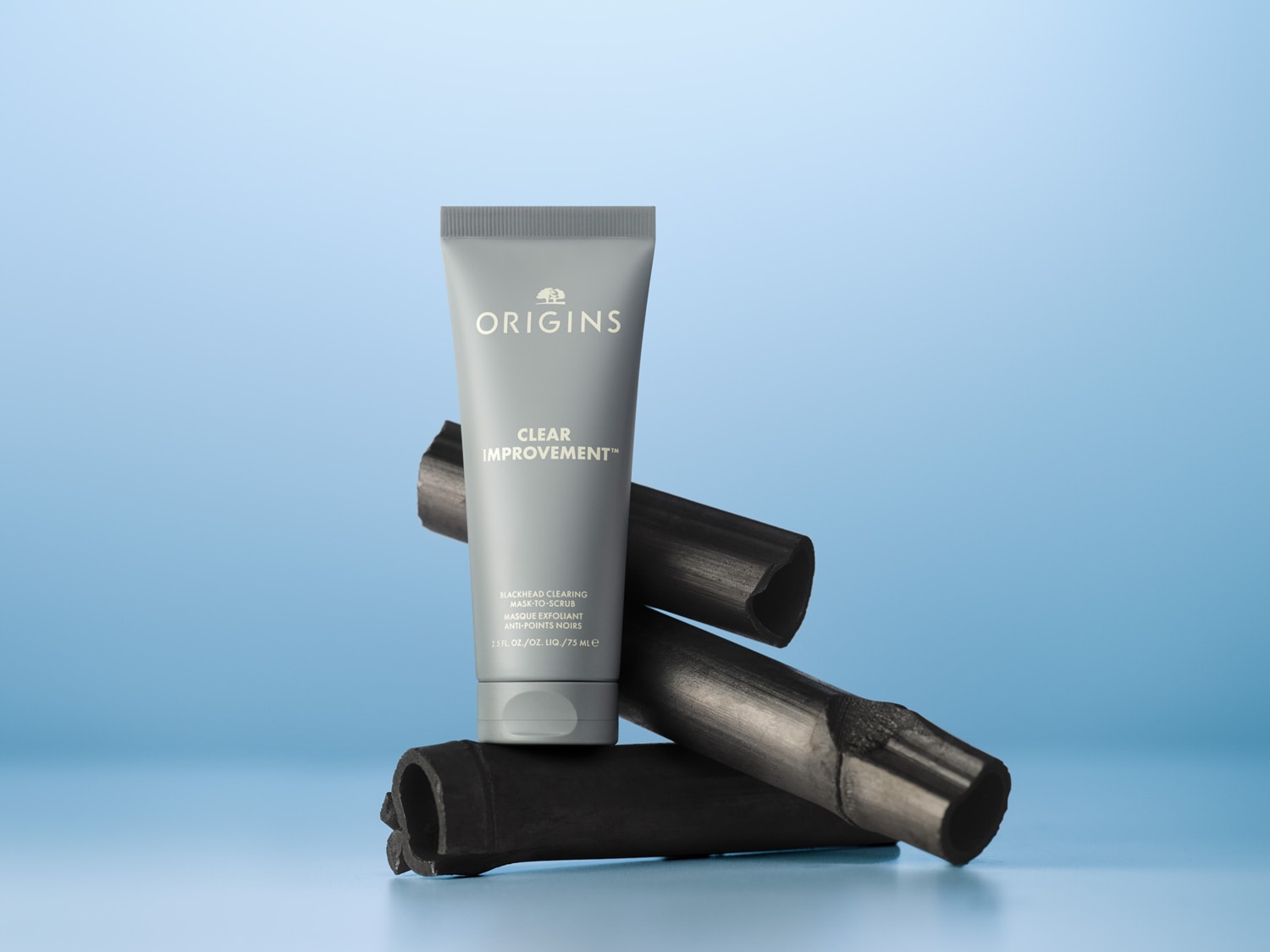 De Origins Clear Improvement masker-to-scrub zit in een grijze tube en staat op stukken houtskool, het masker is voor het reinigen van de huid en het verwijderen van mee-eters.