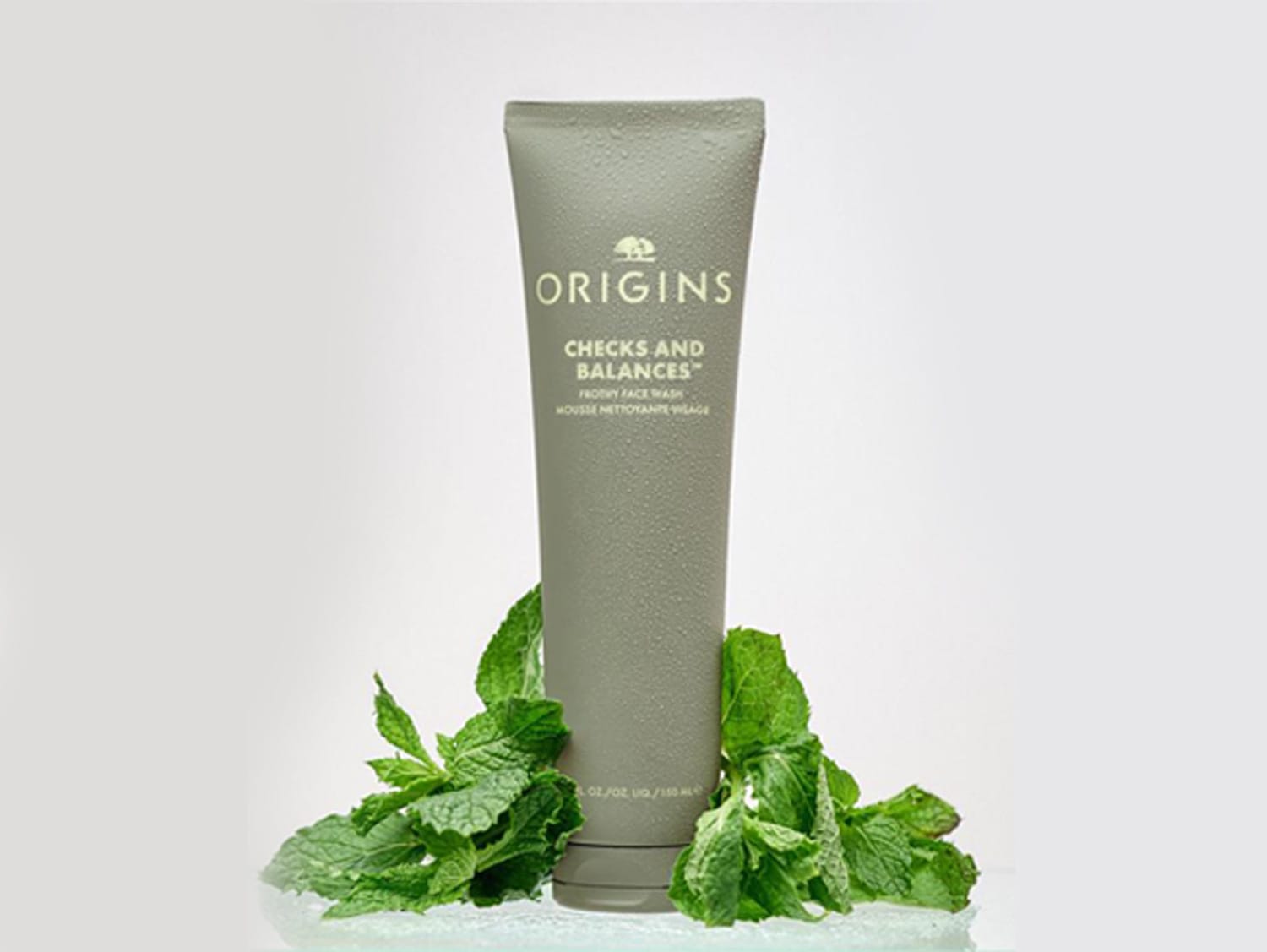 Grijze tube Origins Checks and Balances Frothy Face Wash, omringd door verse muntblaadjes, voor een zachte en evenwichtige reiniging.