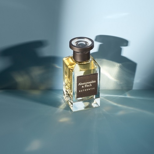 Nahaufnahme eines quadratischen, transparenten Glasflakons mit goldfarbenem Parfüm von Abercrombie & Fitch Authentic, der auf einer glänzenden blauen Oberfläche steht.