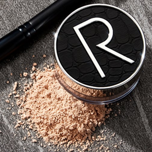 Puder do twarzy w okrągłym, czarnym opakowaniu z srebrną literą R, obok leży pędzel do makijażu i rozsypany puder.