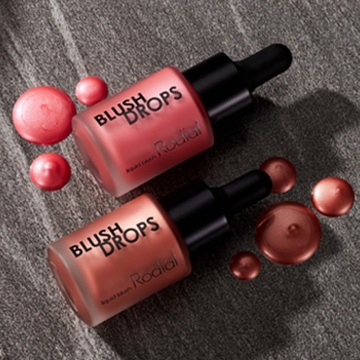 Dwa flakony Rodial Blush Drops z zakraplaczami w odcieniach różu i brązu, otoczone kroplami płynnego różu, prezentują się na szarym tle.