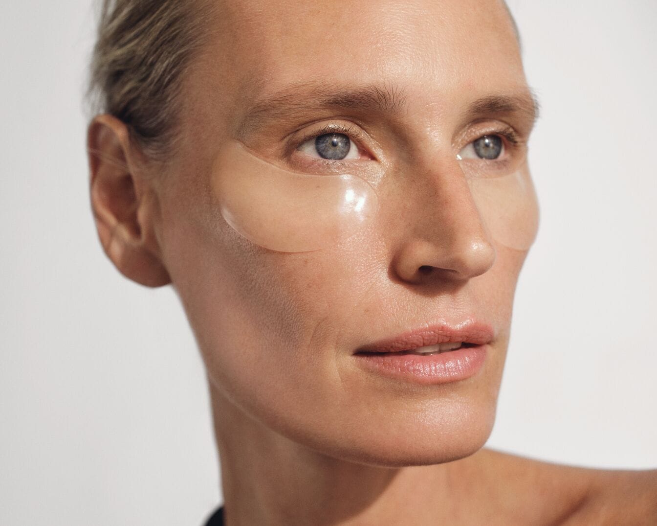 Nahaufnahme einer Frau mit hellblonden, zurückgekämmten Haaren und transparenten Gel-Augenpads unter den Augen, die für einen frischen und strahlenden Blick sorgen.