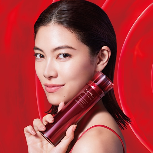 Eine Frau mit glattem, dunklem Haar hält eine rote Shiseido Revitalizing Essence Flasche vor einem roten Hintergrund.