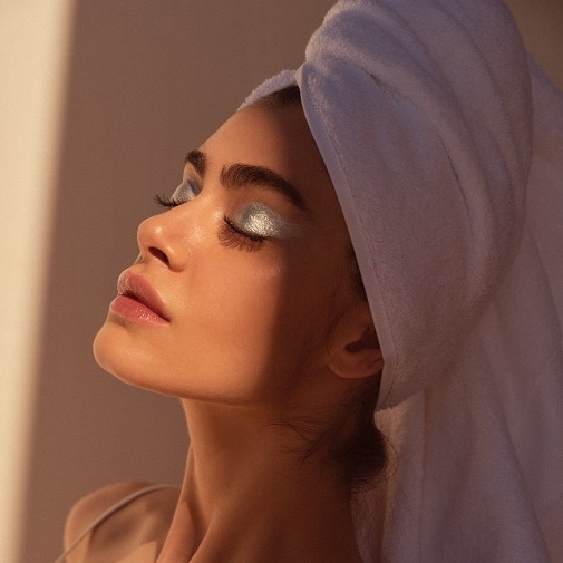 Una mujer con una toalla blanca en la cabeza y sombra de ojos plateada brillante, con la cara iluminada por el sol.