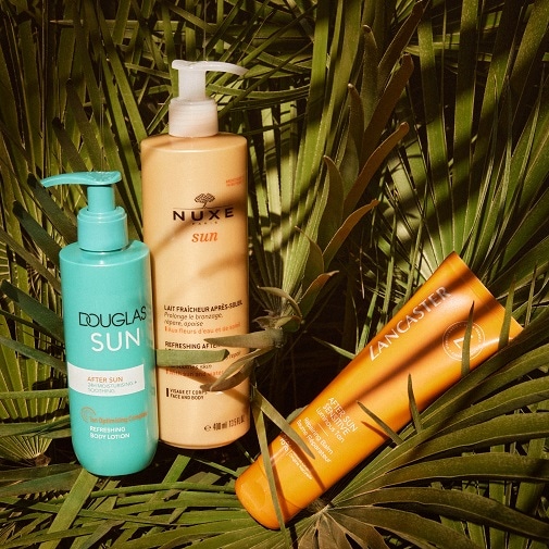 Tres botellas de loción para después del sol de Douglas, Nuxe y Lancaster, con envases en tonos azul, beige y naranja, respectivamente, sobre un fondo de hojas de palma.